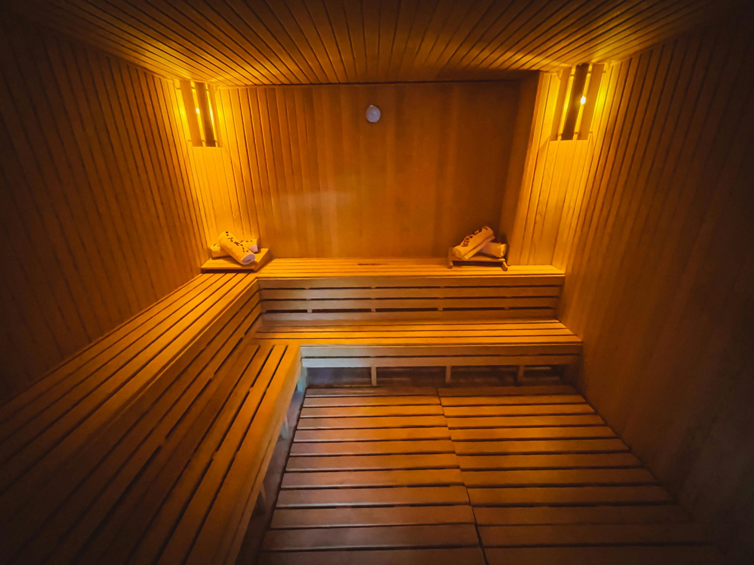 A wooden sauna with two rows of seating positions, we are viewing it at a right angle. There are two sets of towels on the top row, and a light in each corner. There are rows of timber that make up the floor.