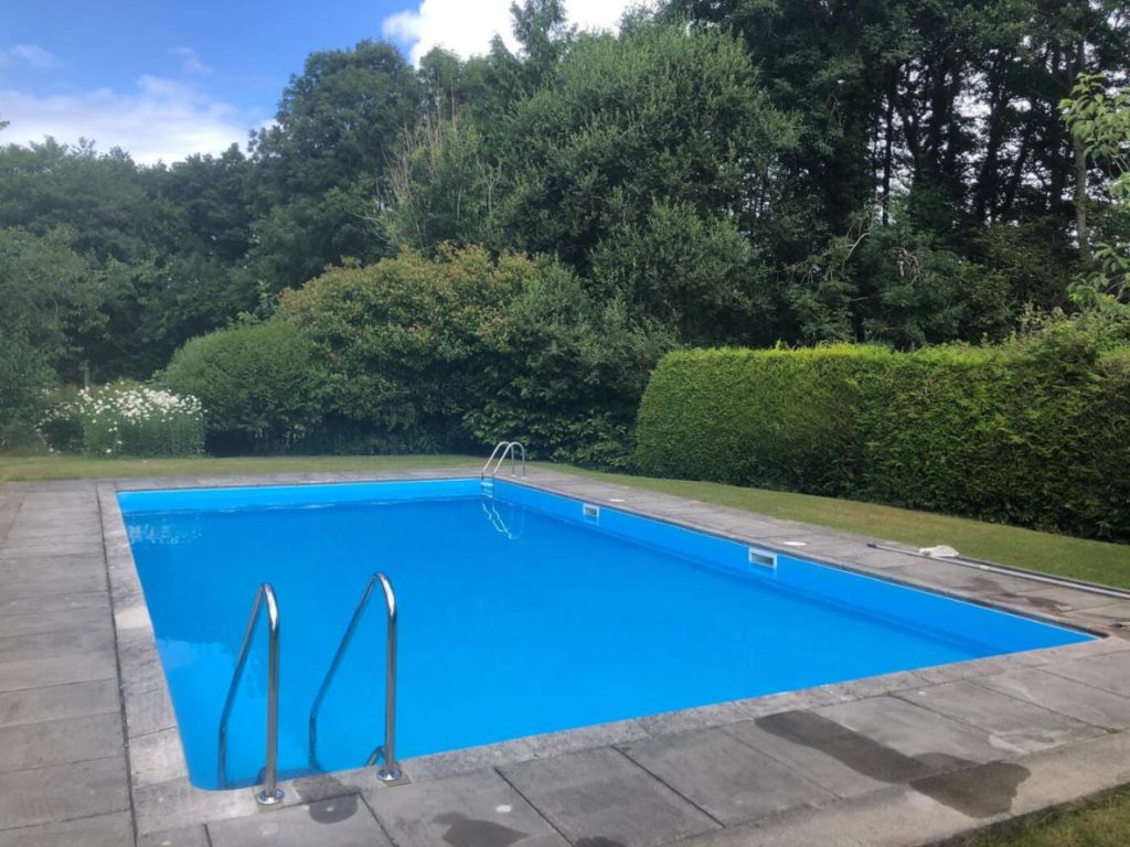 Crystal clear blue water in an empty outdoor swimming pool, surrounded by green grass, lush trees, and hedges on a sunny day. Perfect for summer relaxation.
