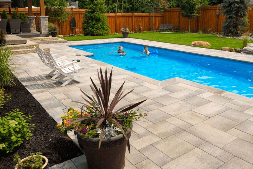 Luxurious backyard with a swimming pool, interlocking stone patio, two kids swimming, and a golden retriever relaxing on the grass
